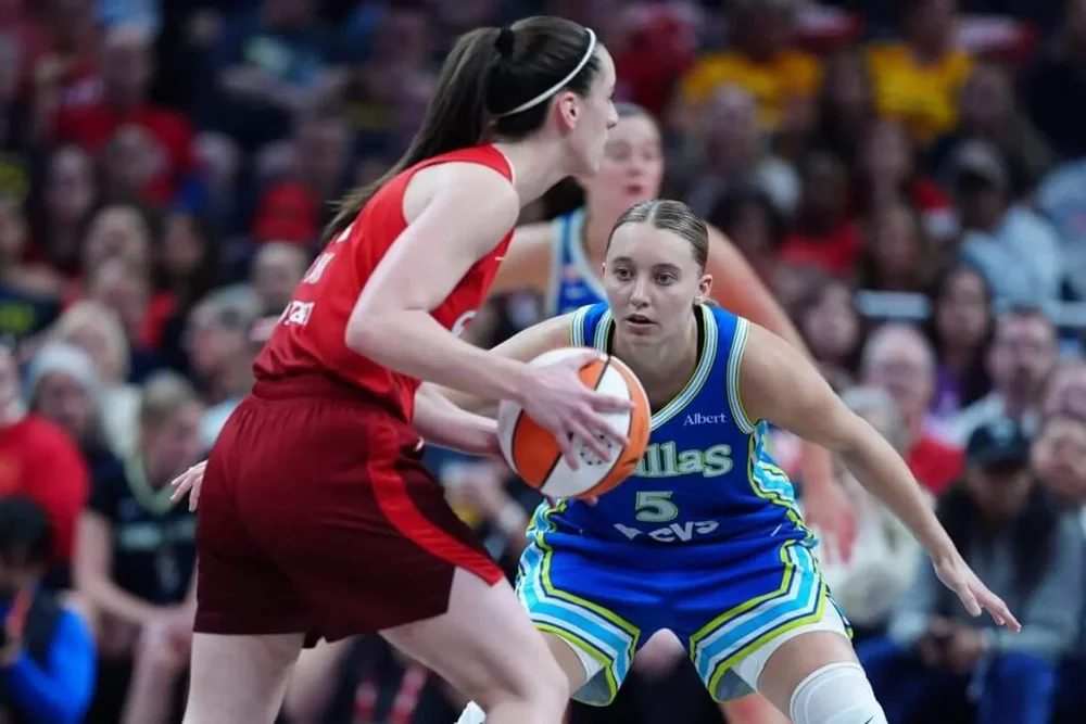 Caitlin Clark holds the ball and looks on as Paige Bueckers guards against her.
